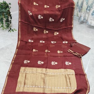 May include: A maroon saree with gold embroidery and a gold border. The saree features a detailed gold pattern at the bottom and small gold floral designs throughout. The saree is folded with a matching piece of fabric.