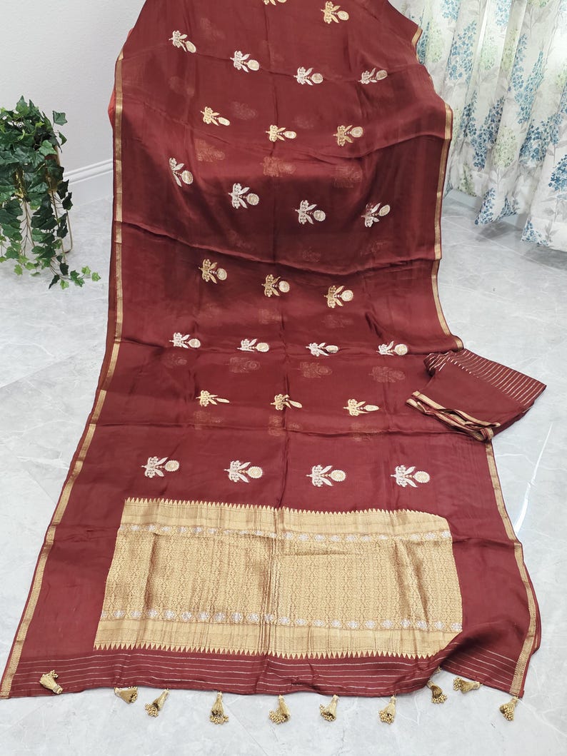 May include: A maroon saree with gold embroidery and a gold border. The saree features a detailed gold pattern at the bottom and small gold floral designs throughout. The saree is folded with a matching piece of fabric.