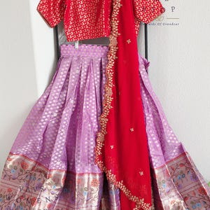 May include: An Indian outfit with a red patterned top and a long, pleated lavender skirt with gold detailing. A red scarf with gold embroidery is draped over the outfit. The skirt has floral and text details.