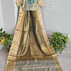 May include: A gold and silver saree with a light blue border and tassels. The saree is draped over a hanger and has a matching blouse with a keyhole neckline. The text "VAVS WOMENS STOP Six Yards of Grandeur" is visible on the image.