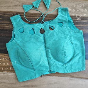 May include: A teal-colored top with cut-out details and a matching tie. The top has a scoop neckline and a fitted silhouette. The fabric has a subtle sheen. The text "VAVS WOMENS STOP" is visible on the left side of the top.