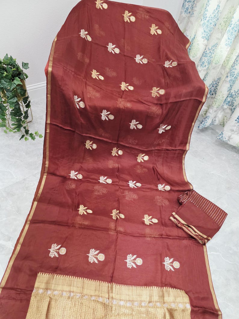 May include: A maroon fabric with gold trim and embroidered floral patterns. The fabric is draped, showcasing the intricate design and texture. A matching rectangular piece is also visible.