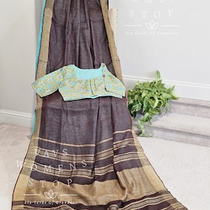 May include: A brown and beige striped linen saree with a blue and gold border. The saree has a matching blue blouse with a floral pattern. The saree is draped over a chair and the text "Vavs Womens Stop Six Yards Of Grandeur" is visible on the wall behind the saree.
