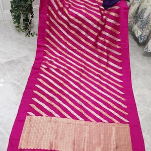 Puede incluir: Un textil magenta vibrante con rayas diagonales blancas y doradas. La tela presenta un borde dorado decorativo en el borde inferior, rematado con borlas magenta. Una tela morada oscura está drapeada sobre el textil.