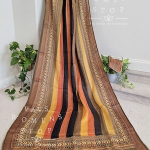 Op de afbeelding: Een bruin, geel, oranje en zwart gestreept stof met een decoratieve rand. De stof heeft een gefranst randje. De tekst "Vavs Womens Stop Six Yards of Grandeur" is op de stof gedrukt.