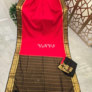 May include: A vibrant red saree featuring a wide black border with intricate gold zari patterns. The pallu is black with horizontal gold stripes and black tassels at the edge. The word VAVS is printed in white in the center of the red fabric.
