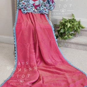 May include: A pink saree with a blue crochet trim. The saree is draped over a chair and has a floral print blouse. The text "Vavs Womens Stop Six Yards Of Grandeur" is visible on the image.