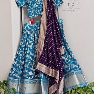 以下が含まれることがあります： ターコイズとシルバーの花柄のスカートとトップのセットで、お揃いの紫と白のドットのスカーフが付いています。セットは黒いハンガーに展示されています。「VAVS WOMENS STOP」の文字が見えます。