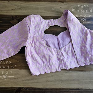 May include: A lavender-colored blouse with a scalloped edge and a keyhole neckline. The blouse has a white floral pattern and the text "Vavs Womens Stop Six Yards Of Grandeur" is printed on the bottom.