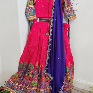 May include: A bright pink Indian outfit, comprising a top, skirt, and a purple scarf. The skirt and sleeves display intricate, colourful patterns with elephants and trees. The scarf is decorated with delicate silver embellishments.