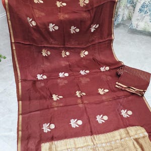 May include: A maroon textile with gold accents and embroidered floral patterns. The fabric has a sheer quality, with a gold border and a matching piece of fabric. The design features repeating floral motifs in white and gold.