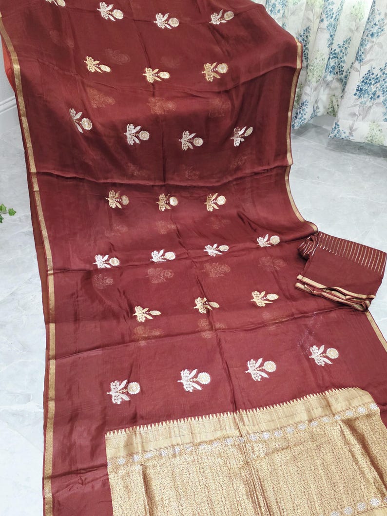 May include: A maroon textile with gold accents and embroidered floral patterns. The fabric has a sheer quality, with a gold border and a matching piece of fabric. The design features repeating floral motifs in white and gold.