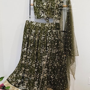 May include: An olive green two-piece outfit with a crop top and a long skirt, embellished with white and gold accents. The top has a square neckline and a tie closure. The skirt has a wide hem with gold trim. A matching sheer scarf is draped on the side.