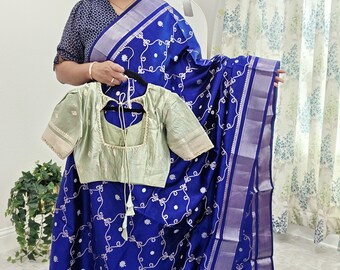 May include: A woman holding a blue saree with white floral embroidery and a light green blouse. The saree has a silver border and a matching blouse with short sleeves. The saree and blouse are traditional Indian garments.