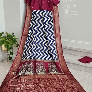 Op de afbeelding: Een traditionele Indiase outfit met een bordeauxrode blouse met pofmouwen en een wit en marine chevron patroon rok. De set bevat een bordeauxrode sjaal met ingewikkelde gouden en witte details en een bijpassend stuk stof.