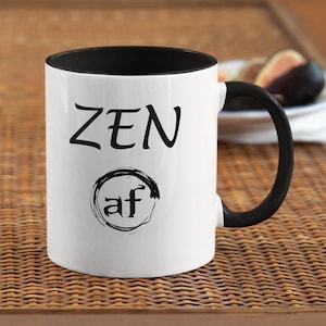 May include: White ceramic mug with a black rim and handle. The mug has black text that reads "ZEN af" with a black circle around the "af" and a black brushstroke design.
