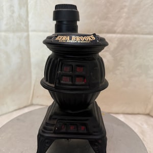 May include: Black ceramic bottle shaped like a wood-burning stove with a gold label that reads "Ezra Brooks Real Sippin' Whiskey".