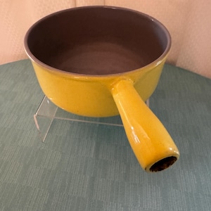 May include: A yellow enamel saucepan with a single handle. The interior is a dark gray color, contrasting with the bright exterior. The pan is sitting on a clear acrylic stand.
