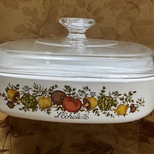 May include: White oval casserole dish with a glass lid. The dish has a floral design with a border of green leaves, red and yellow fruits, and brown mushrooms. The text "L'Echalote" is printed on the bottom of the dish.