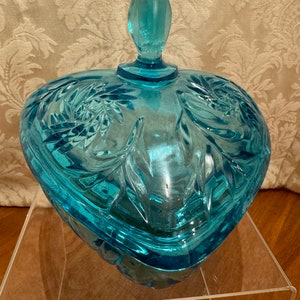 May include: A heart-shaped glass candy dish with a lid. The dish is made of clear blue glass and has a floral design. The lid has a knob on top.