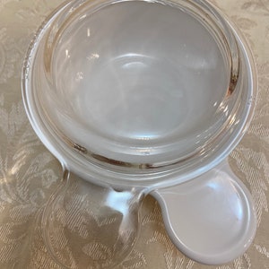 May include: A clear glass Pyrex bowl with a white handle on each side. The bowl is round and has a wide rim. The handles are petal-shaped. The bowl is sitting on a light-colored surface.