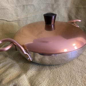 May include: A copper and stainless steel casserole dish with a black knob on the lid. The dish has two handles on the sides.