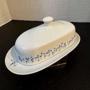 May include: White ceramic butter dish with a lid and a matching oval-shaped base. The dish is decorated with a delicate blue floral pattern around the edges. The lid has a small, round knob for easy lifting. A classic piece for the kitchen.