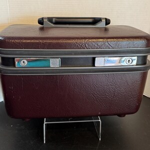 May include: A burgundy vintage suitcase with a textured exterior, a black handle, and silver-colored latches. The suitcase is rectangular and sits on a clear plastic stand. The suitcase is a travel accessory.