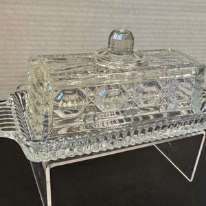 May include: A clear, rectangular glass butter dish with a lid. The dish has a decorative pattern of geometric shapes and a handle on the lid. The base of the dish has a ribbed design. The butter dish is displayed on a clear stand.