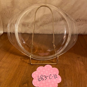 May include: A clear glass Pyrex baking dish with a lid, resting on a gold-colored metal stand. The dish is round with handles. A pink, flower-shaped tag with the text "683-C-12" is on the wooden surface.