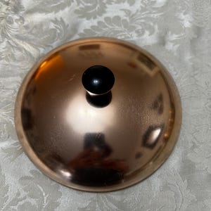 May include: A copper-colored pot lid with a black knob on top. The lid is round and has a shiny finish.