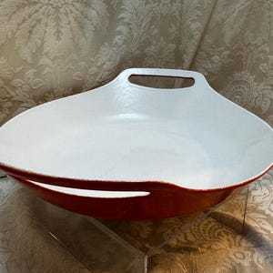 May include: A white and red ceramic serving dish with a handle. The dish has a wide, oval shape with a white interior and a red exterior. The handle is a cutout at the top of the dish. The dish is approximately 14 inches long.