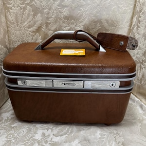 May include: A vintage brown Samsonite suitcase with a silver handle and trim. The suitcase has a rectangular shape with rounded corners and a leather luggage tag attached. The Samsonite logo is visible on the front.