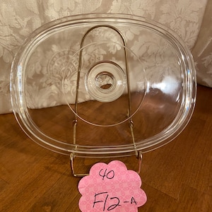 May include: A clear glass lid for a slow cooker with a metal handle. The lid is sitting on a wooden surface with a pink note that says "40 F12-A".