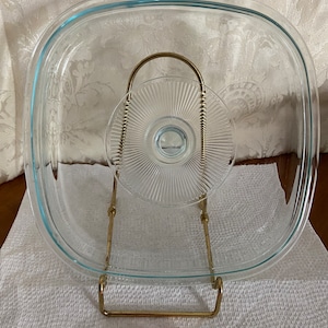 May include: A clear glass baking dish with a gold metal stand. The dish is round and has a slightly raised edge. The stand has a round top and three legs.