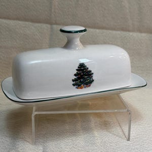 May include: White ceramic butter dish with a green trim and a decorative Christmas tree design. The lid has a small handle with a green accent. The dish sits on a clear acrylic stand.