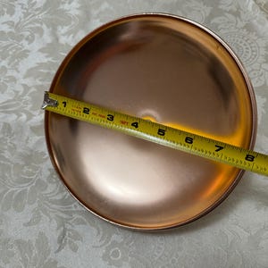 May include: A copper-colored metal cake plate with a diameter of 8 inches. The plate is round and has a slightly raised edge.