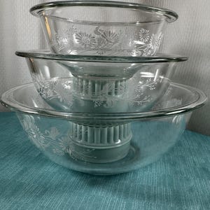 May include: A stack of three clear glass bowls with a floral design. The bowls are nested, with a light green juicer component visible in the center. The bowls are on a teal surface.