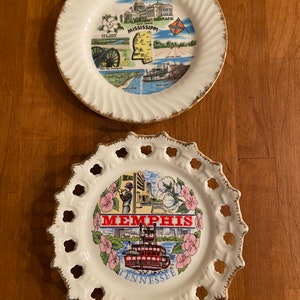 May include: Two vintage souvenir plates. The top plate is white with a gold rim and features a map of Mississippi with various images of the state. The bottom plate is white with a gold rim and a lace design around the edge. It features a colorful image of Memphis, Tennessee with a steamboat and the text "Memphis Tennessee".