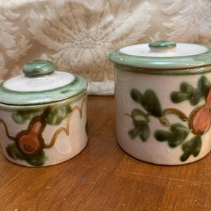 May include: Two ceramic jars with lids, both featuring a green, brown, and white floral design with a pear motif. The larger jar is taller and has a wider opening than the smaller jar.