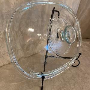 May include: A clear glass oval-shaped lid with a glass knob on top. The lid is sitting on a black metal stand.