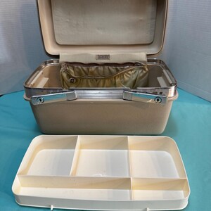 May include: A beige hard-shell train case with a metallic silver handle and a hinged lid. The interior features a beige fabric lining with a zippered pocket and a removable white plastic tray with five distinct compartments for organization.