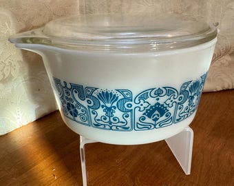 Op de afbeelding: Witte keramische ovenschaal met een blauw-wit geometrisch patroon. De schaal heeft een glazen deksel en staat op een heldere acryl standaard.