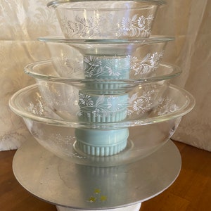 May include: A stack of four clear glass mixing bowls with a white floral design. The bowls are nested on top of a light blue pedestal and a silver metal stand. The stand is on a white base.