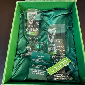 May include: Two clear glass Irish coffee mugs with handles, displayed in a green gift box with green satin fabric. Each mug has the words "Irish Coffee" and a shamrock design. A green tag reads "eamon".