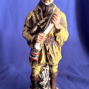 May include: A ceramic figurine of a man in a brown hat and yellow coat holding a rifle. The figurine is standing on a tree stump and has a label on the base that reads "Beam 1890 Kentucky Straight Bourbon Whiskey 80 Proof".