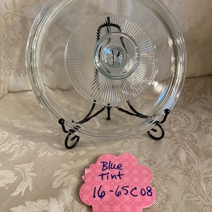 May include: A clear glass lid with a blue tint and a raised center knob. The lid is sitting on a black metal stand. A pink note with the text "Blue Tint 16-65C08" is in the foreground.