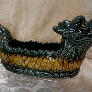 May include: A green and brown ceramic dragon planter with a red accent on the snout. The dragon is shaped like a boat with a wide, open mouth.