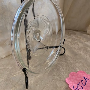 May include: Clear glass lid with a raised, circular pattern in the center. The lid is on a black metal stand.