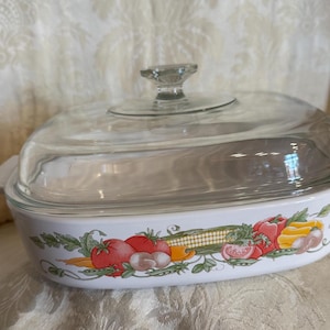 May include: White ceramic baking dish with a glass lid. The dish has a floral design with vegetables and a corn cob.